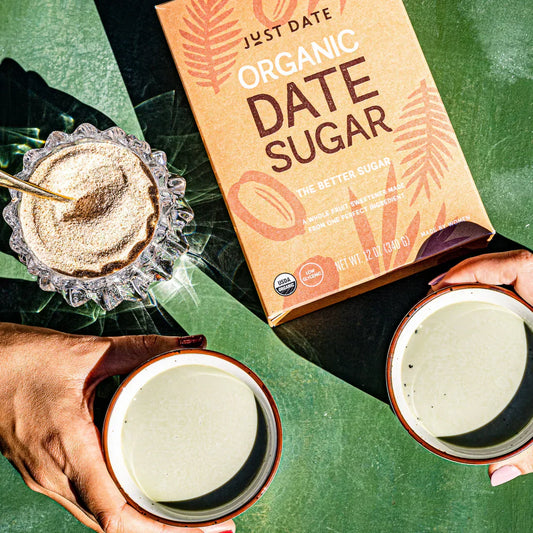 Hands holding lattes beside a Just Date Organic Date Sugar box and a bowl of date sugar on a green tabletop.