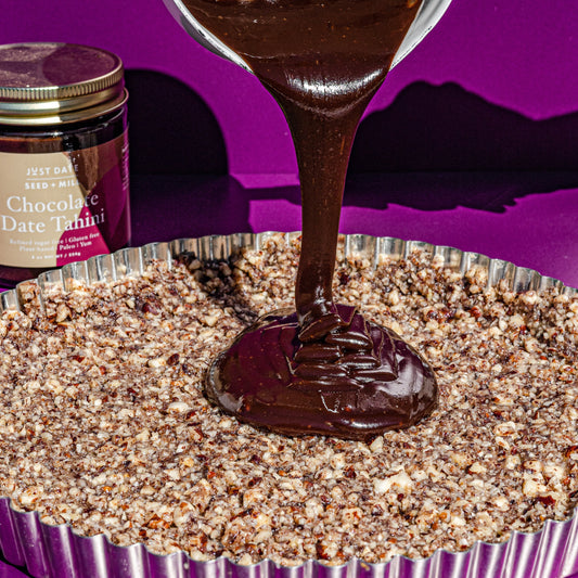 Chocolate ganache being poured from a glass measuring cup into a pecan tart crust, beside a jar of Just Date Chocolate Date Tahini