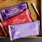 Three mini chocolate chip bags from Just Date are arranged in a wooden tray. The packages are purple (Organic Dark Chocolate Chips), red (Organic Milk Chocolate Chips), and lavender (Date Sweetened Organic Dark Chocolate Chips), with a pink and white marker on a dark gray surface next to the tray.