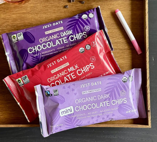 Three mini chocolate chip bags from Just Date are arranged in a wooden tray. The packages are purple (Organic Dark Chocolate Chips), red (Organic Milk Chocolate Chips), and lavender (Date Sweetened Organic Dark Chocolate Chips), with a pink and white marker on a dark gray surface next to the tray.
